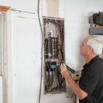 Un homme examine un tableau électrique avec des outils dans une pièce blanche