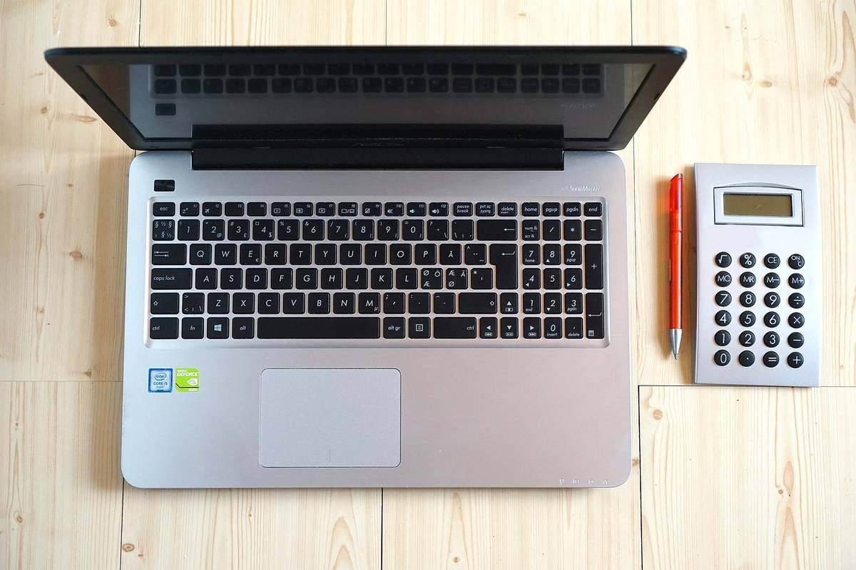 Ordinateur portable gris ouvert à côté d'une calculatrice de bureau blanche et d'un stylo orange sur bois Ordinateur portable gris ouvert à côté d'une calculatrice de bureau blanche et d'un stylo orange sur bois
