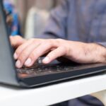 Main d'une personne tapant sur le clavier d'un ordinateur portable noir posé sur un bureau blanc