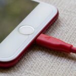 Smartphone blanc avec câble de charge rouge branché, posé sur textile beige
